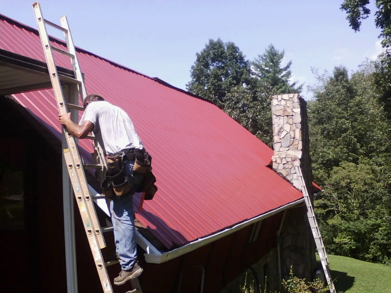 Roofer on a ladder beside a red metal roof and stone chimney — Soffit & Fascia Repair by Roofing Brothers in Meadow Valley